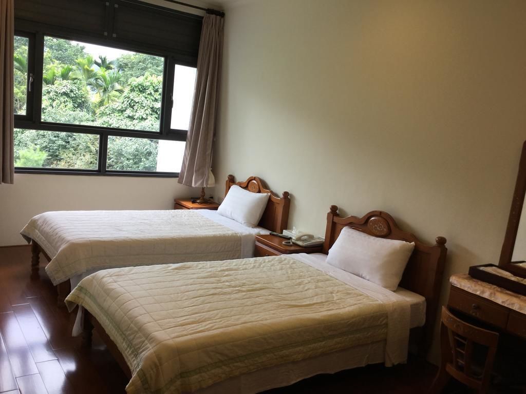 A room with two beds, a window with a view of greenery, a wooden nightstand, and a desk with a mirror.