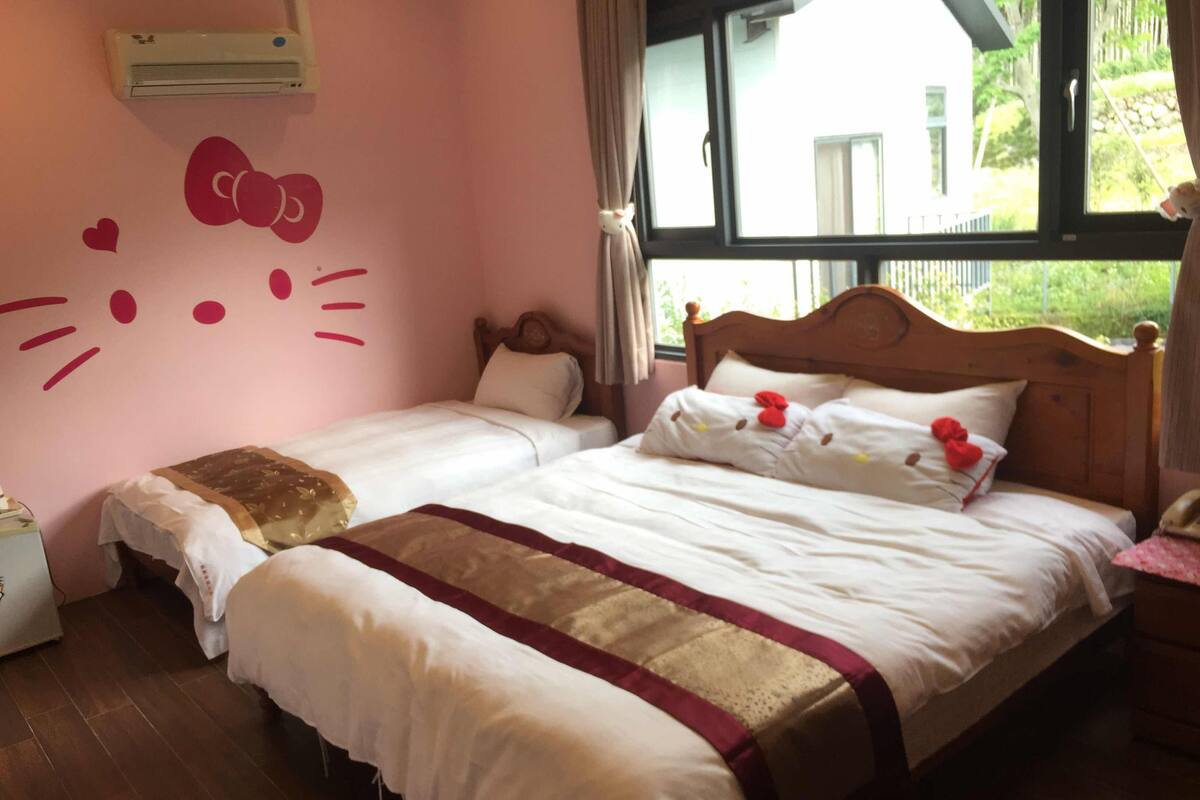 Une chambre avec deux lits simples, une grande fenêtre et un autocollant mural à l’effigie de Hello Kitty.