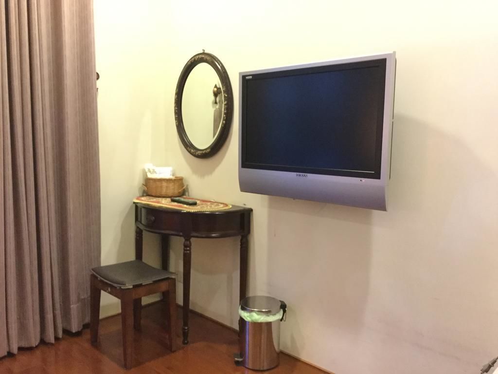 A room with a flat-screen TV mounted on the wall, a small wooden table with a basket and a tissue box, a wooden chair, and a trash can.