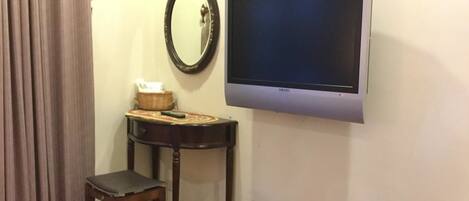A room with a flat-screen TV mounted on the wall, a small wooden table with a basket and a tissue box, a wooden chair, and a trash can.