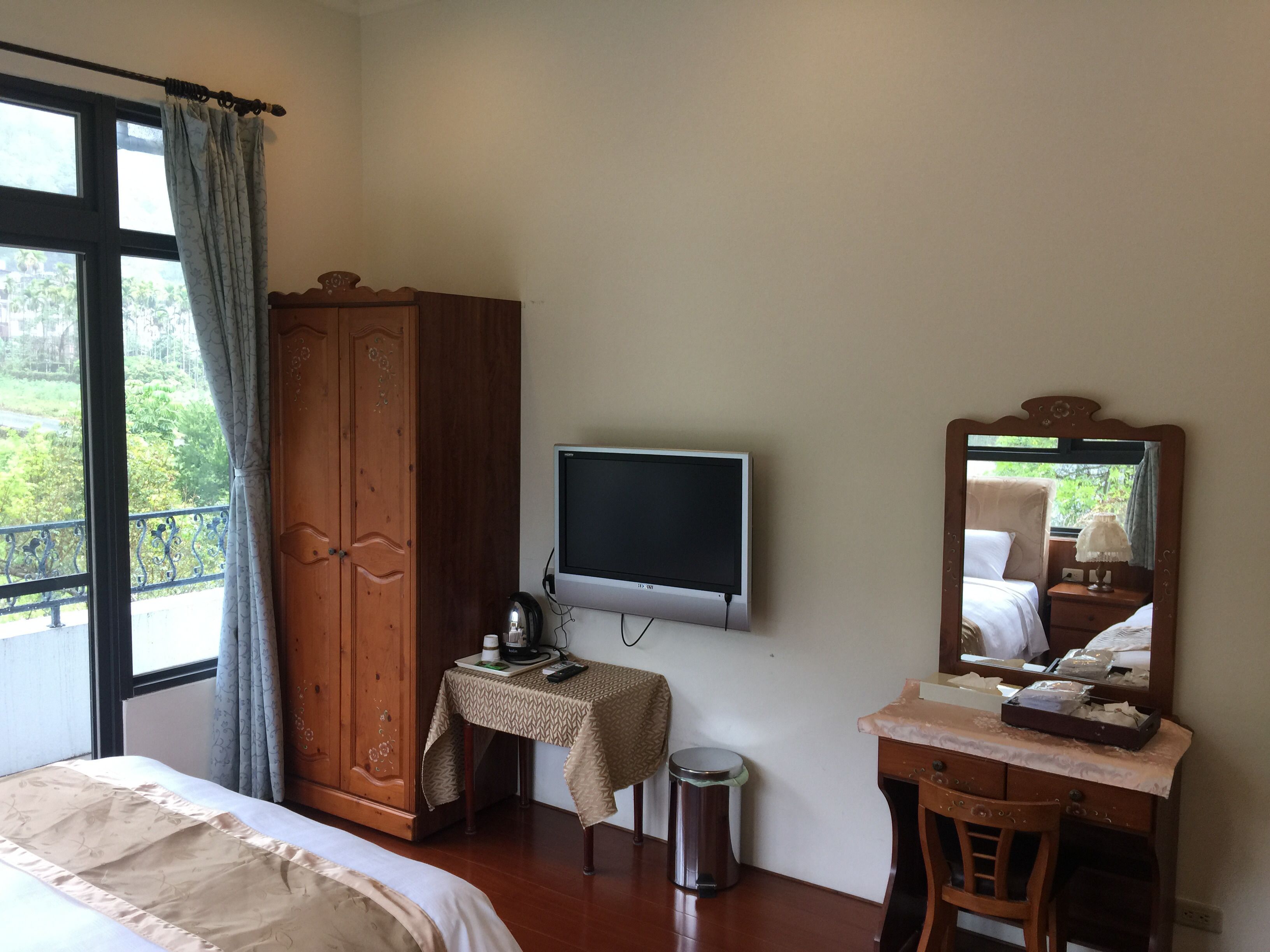 A bedroom with a bed, a wooden wardrobe, a television, a small table with a lamp, and a trash bin.