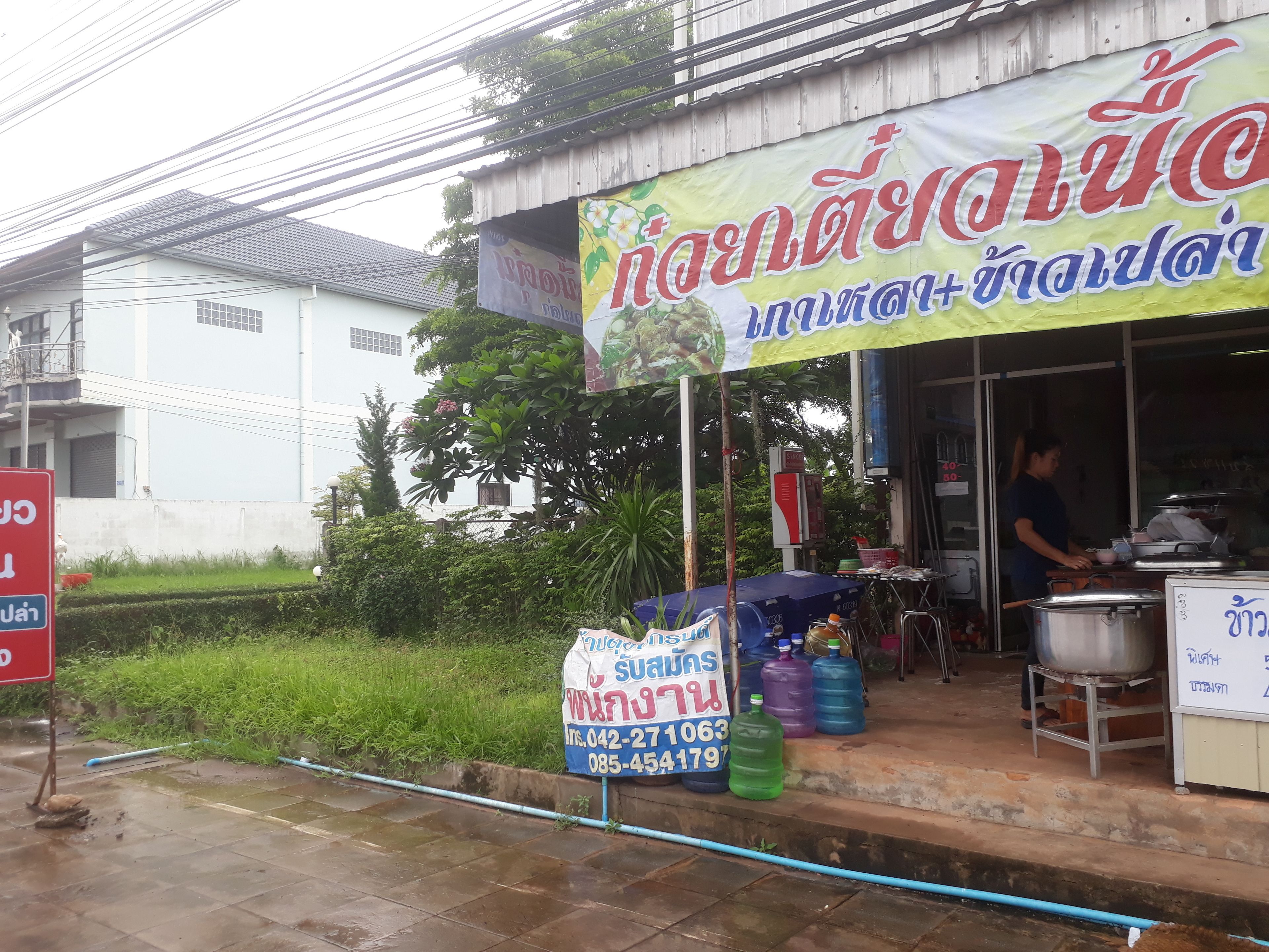 A roadside stall with a large sign in a foreign language, water dispensers, and a person cooking inside.