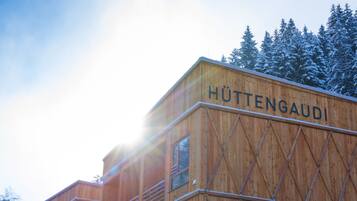 Una cabaña de madera con el nombre 'HÜTTENGAUDI' en su lateral, rodeada de árboles cubiertos de nieve.