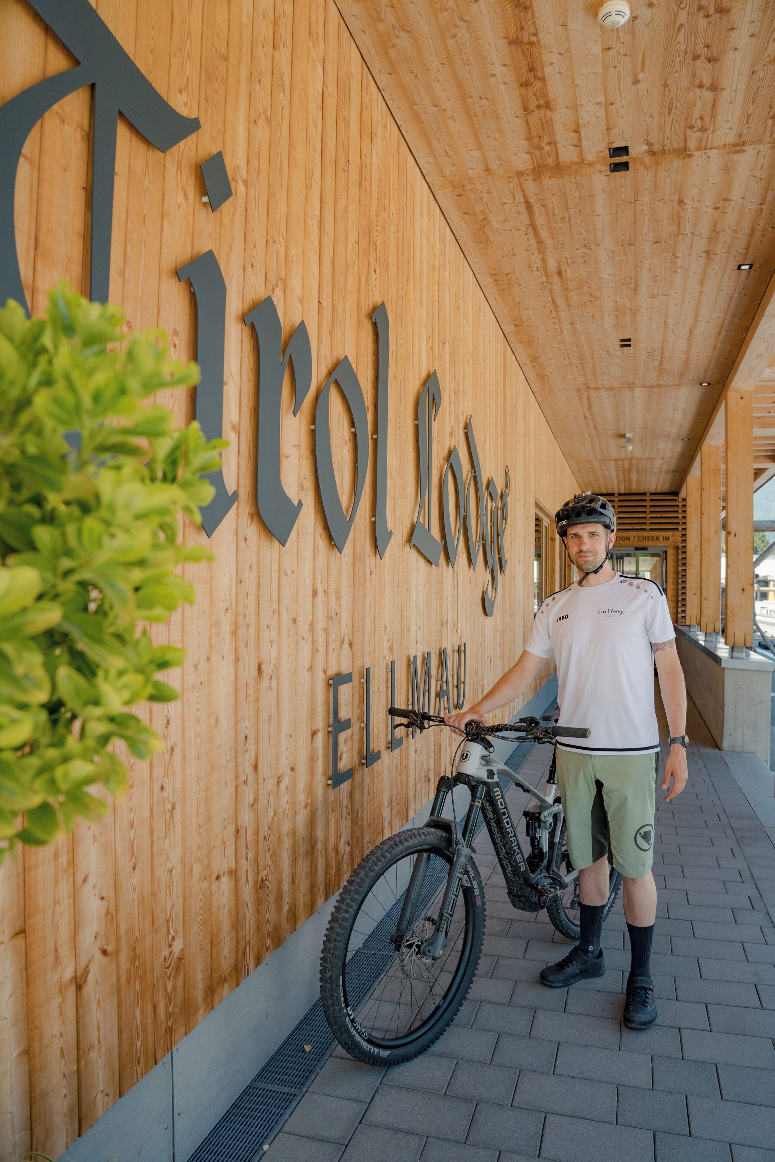 A cyclist standing next to a mountain bike in front of a wooden wall with the text 'Trolodge ELLMAU'.