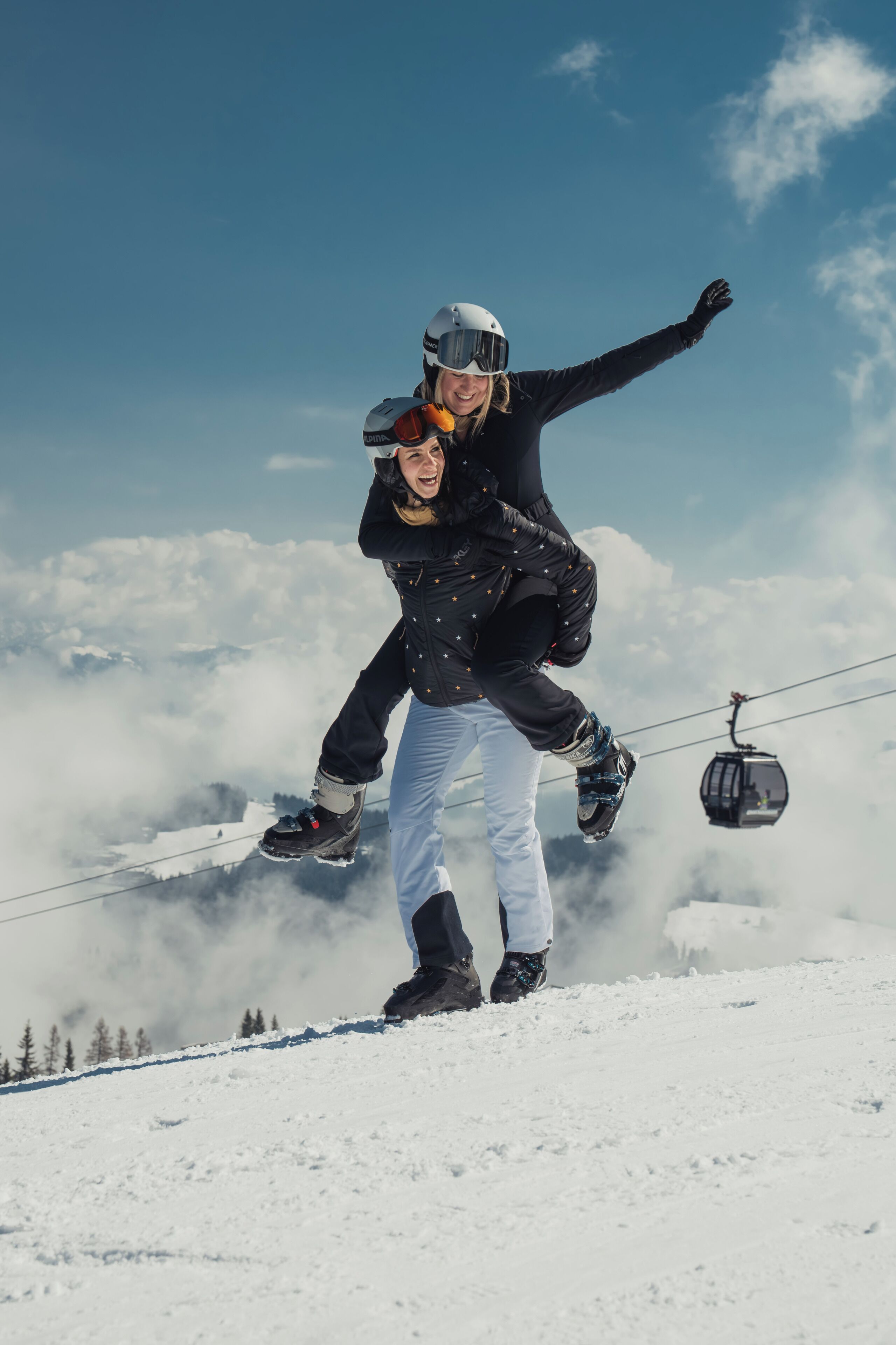 Una persona con chaqueta negra y pantalones blancos en una montaña nevada, con un teleférico al fondo.