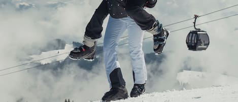 Una persona con chaqueta negra y pantalones blancos en una montaña nevada, con un teleférico al fondo.