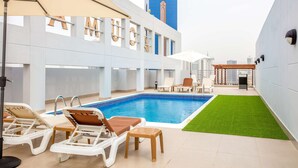 A rooftop pool area with lounge chairs and umbrellas, a view of a tall building in the background, and a green artificial turf area. - Loumage S Suites and Spa (Manama)