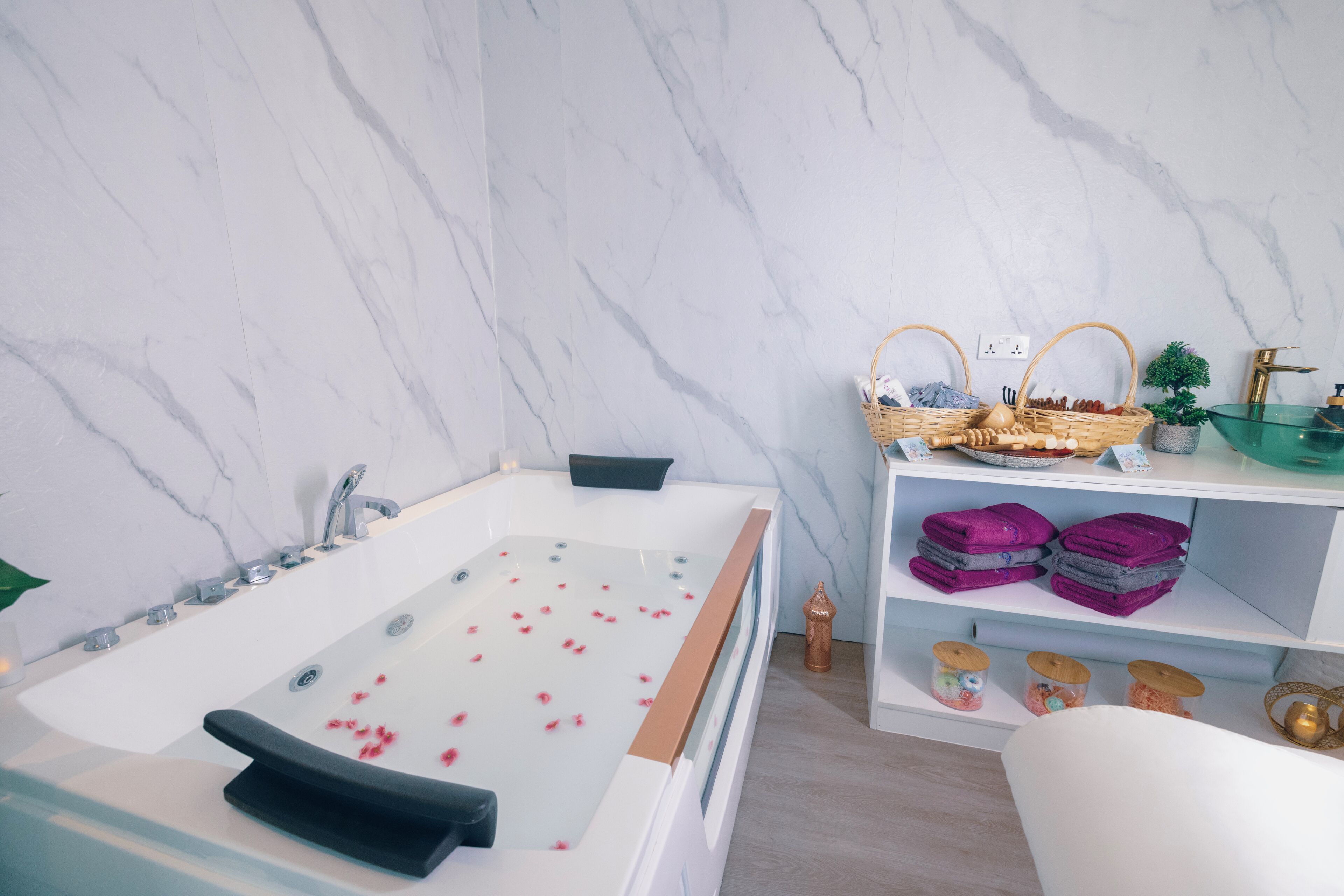 A modern bathroom with a marble wall, a bathtub with rose petals, a shelf with towels, and a small plant.