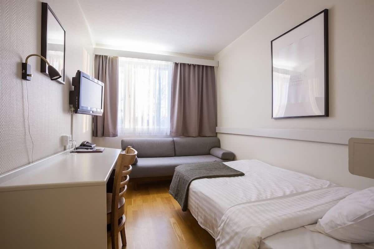 Ein Hotelzimmer mit Bett, Schreibtisch, Stuhl, Fernseher, Fenster mit Vorhängen und einem gerahmten Bild an der Wand.