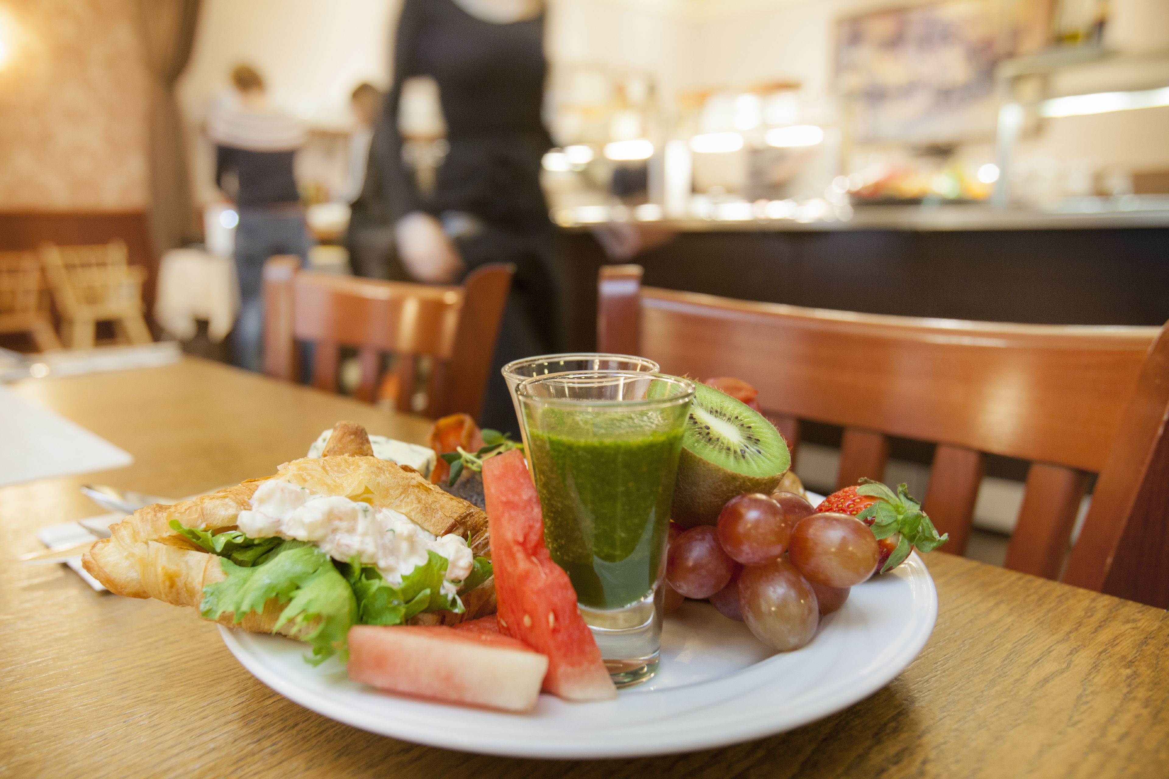 En tallerken med en sandwich, frugt og et glas grøn juice på et træbord i en restaurant.