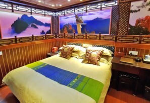 A traditional-style hotel room with a large bed, decorative pillows, a nightstand with a lamp, and a wall-mounted TV displaying a scenic view. - Yangshuo Li River Gallery Lodge (Guilin)