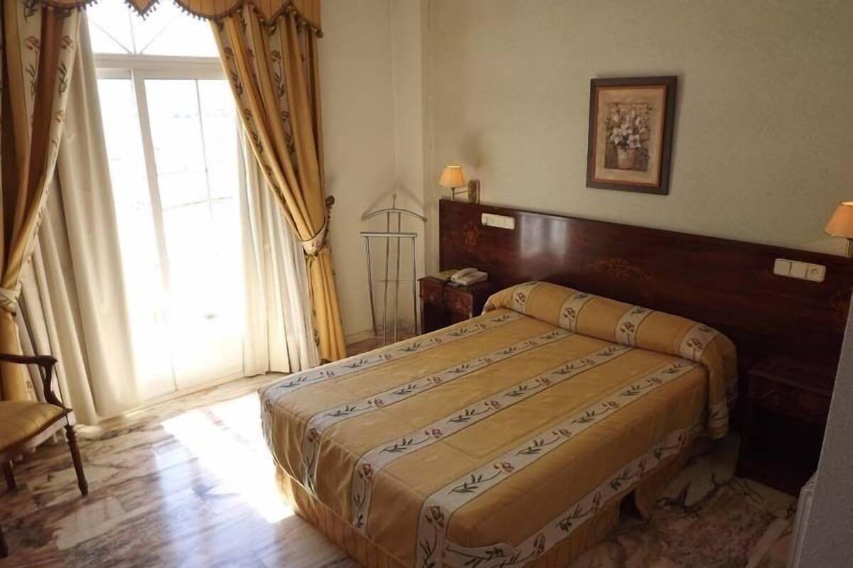 A hotel room with a bed, a chair, a bedside table, a lamp, and a window with curtains.