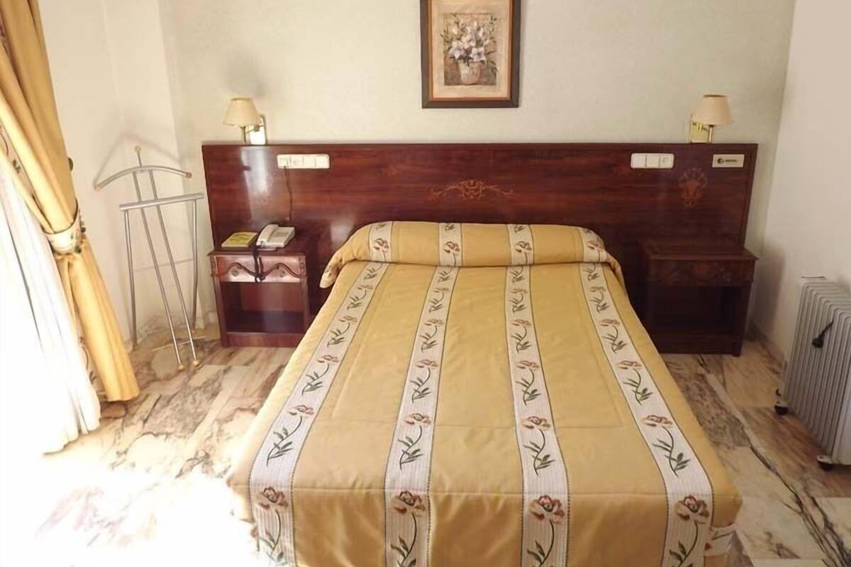 A hotel room with a wooden headboard, a bed with a floral bedspread, a nightstand with a lamp, and a radiator.