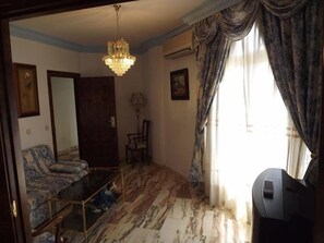 Quarto com lustre, sofá, poltrona, mesa e uma janela grande com cortinas.