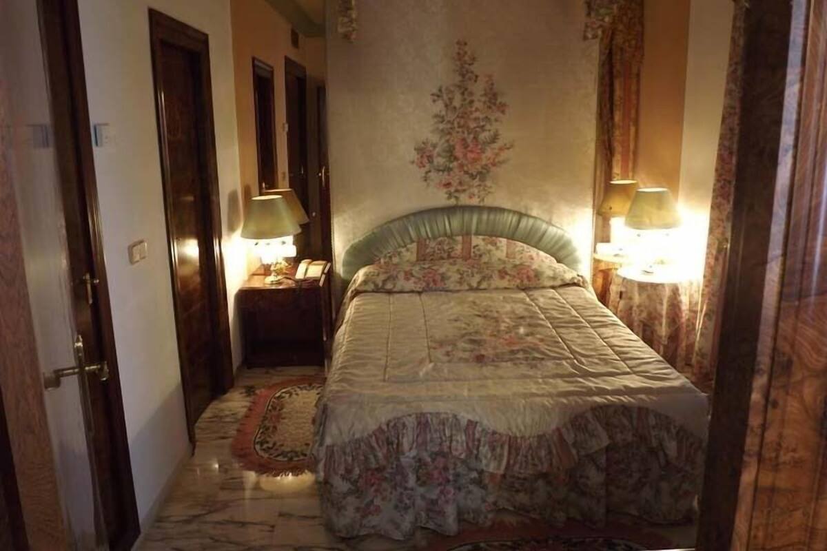 A bedroom with a bed, two bedside lamps, a floral wall decoration, and a patterned rug.