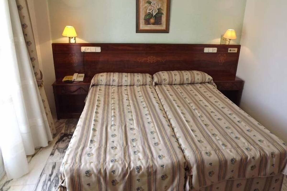 A hotel room with a bed, two bedside lamps, a nightstand, a framed picture on the wall, and a curtain.