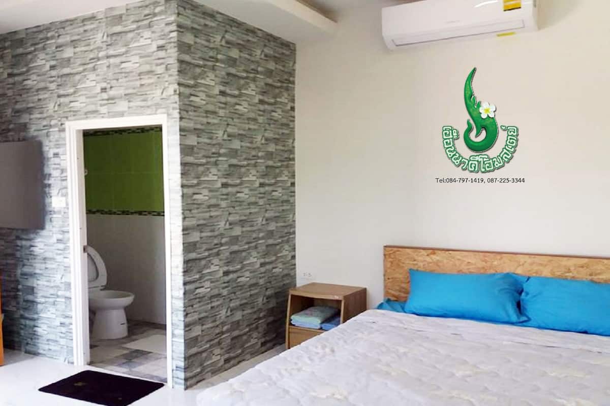A bedroom with a bed, a nightstand, a wall-mounted air conditioner, and a bathroom with a toilet and shower.