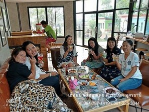 A group of people sitting around a table with food and drinks, in a room with large windows and a view of the outside. - Heuan Na Khee (Ban Dung)