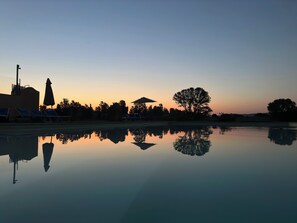Piscina tranquila com cadeiras de descanso e guarda-sóis, cercada por árvores e edifícios ao pôr do sol.