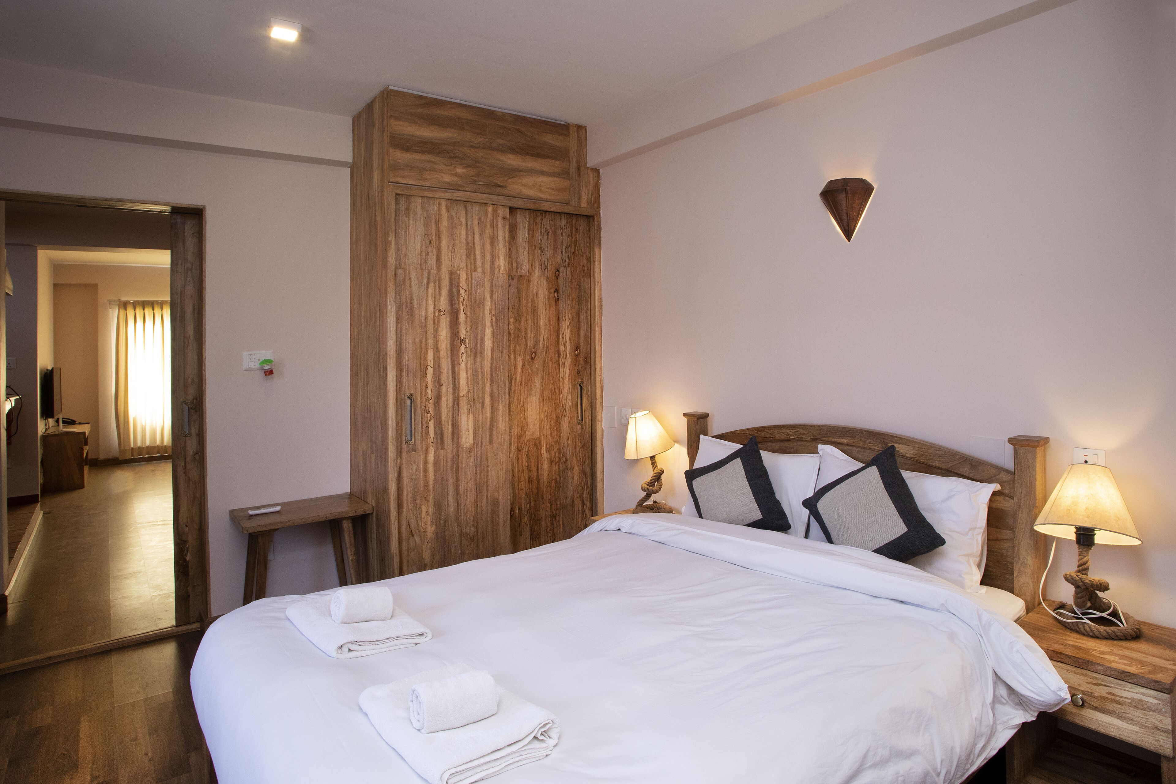 A neatly made bed with white linens and towels, a wooden wardrobe, a bedside lamp, and a wall-mounted light fixture.