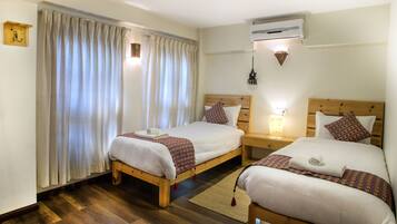Habitación de hotel con dos camas, suelo de madera, un ventanal grande con cortinas, un equipo de aire acondicionado y una lámpara de noche.