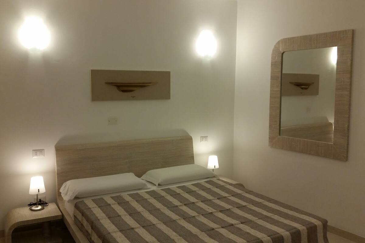 Une chambre d’hôtel avec un lit, deux lampes de chevet, un miroir fixé au mur et une petite étagère au-dessus du lit.