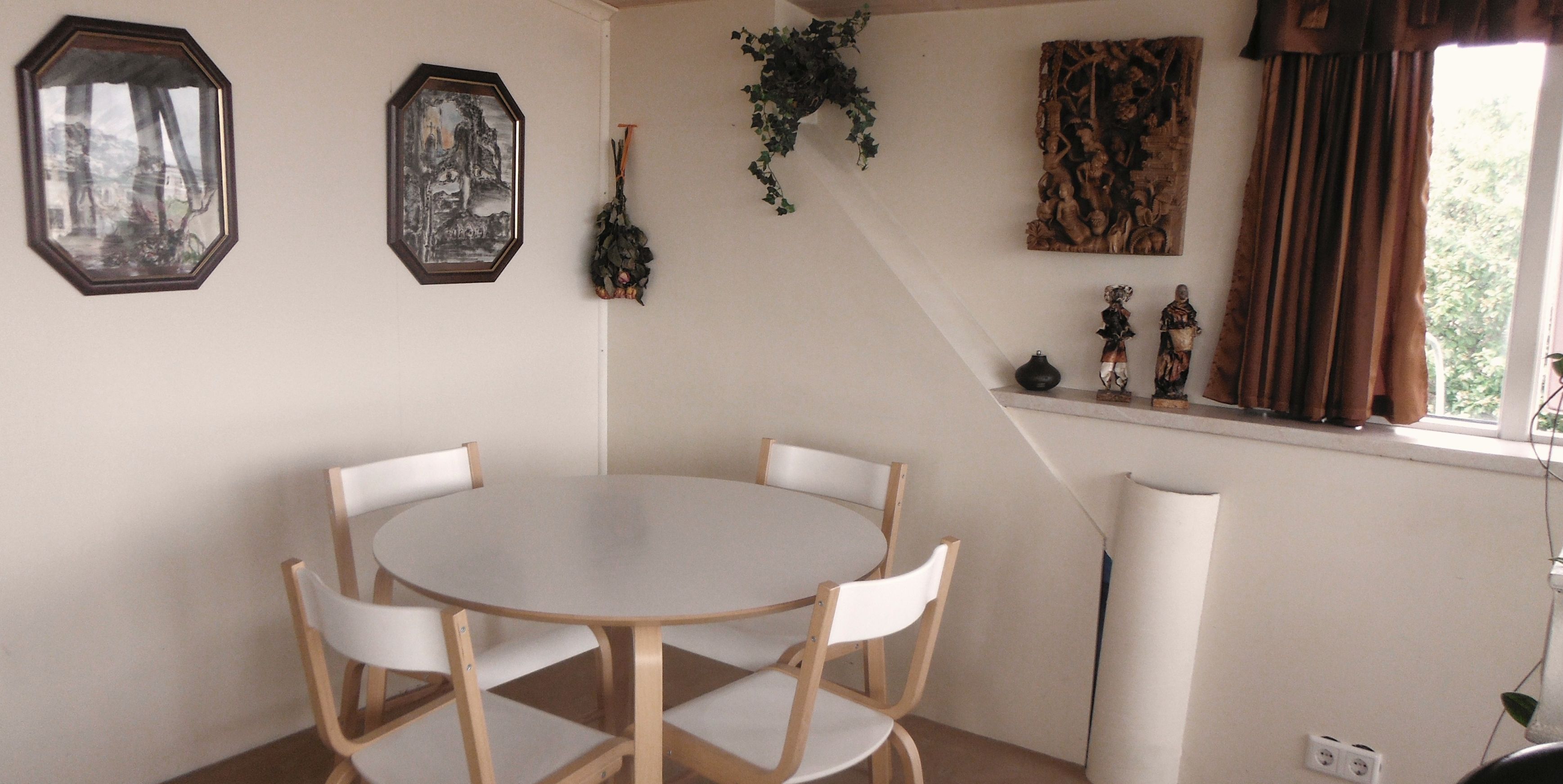 A dining area with a round table and four chairs, a window with curtains, and wall decorations including framed pictures and a hanging plant.