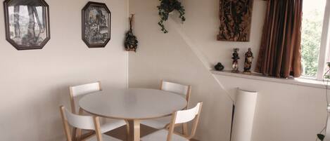 A dining area with a round table and four chairs, a window with curtains, and wall decorations including framed pictures and a hanging plant.