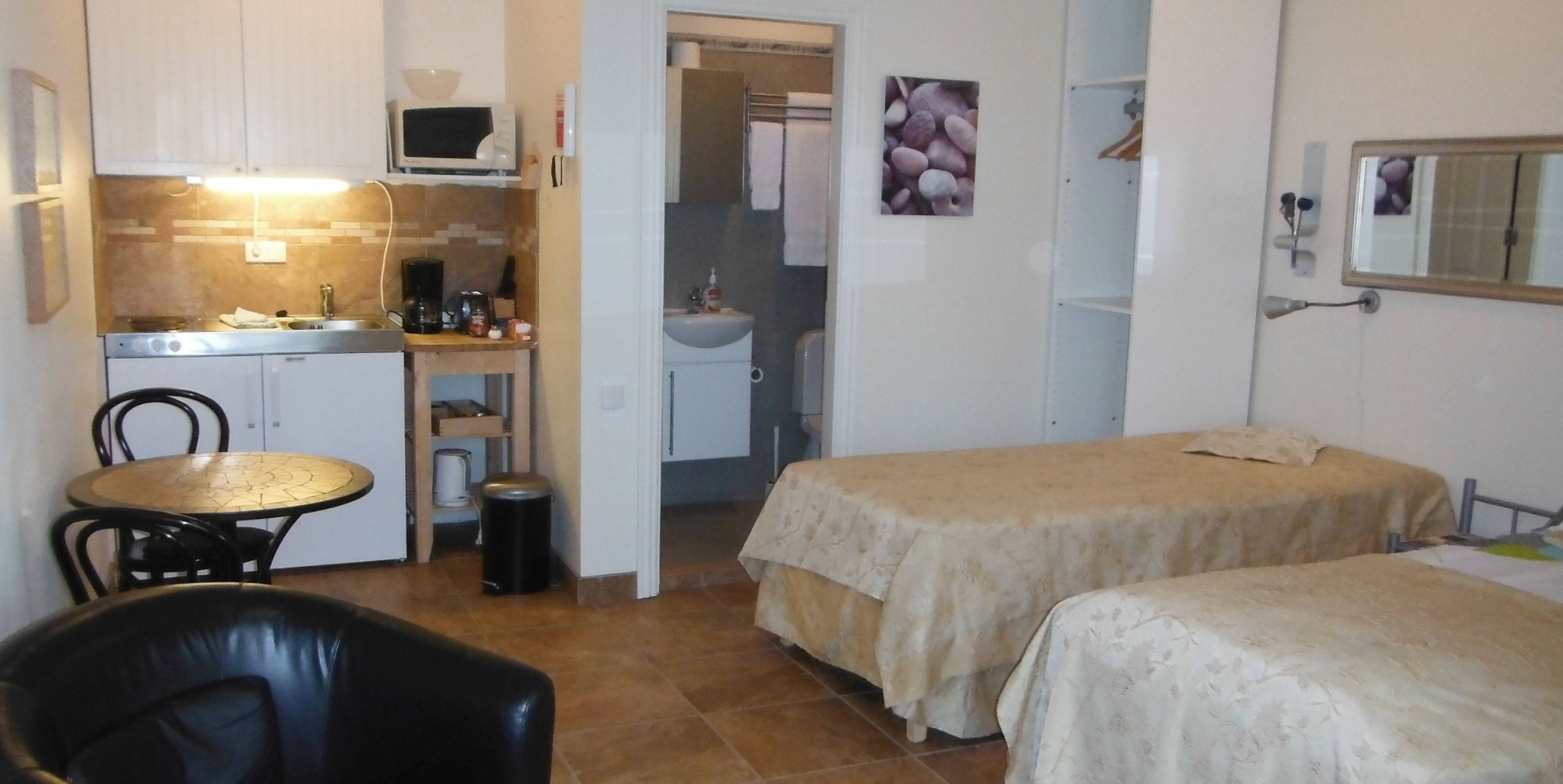 A compact hotel room with a kitchenette, a small dining table, two beds, a bathroom, and a mirror.