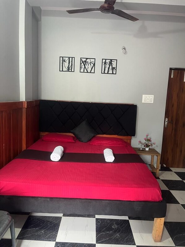 A bed with a black headboard and red bedding, a small bedside table with a flower, and a ceiling fan. - Casa Almeida Guest House (Candolim)