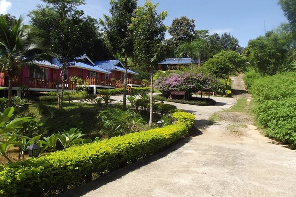 A pathway leading to a row of red-roofed buildings surrounded by lush greenery and a purple flowering bush.
