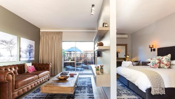 A modern hotel room with a leather sofa, a wooden coffee table, a large bed with a dark headboard, and a balcony with a view.
