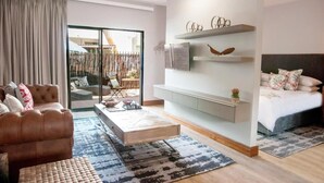 A modern living room with a sofa, coffee table, and a bed with a headboard. There are shelves with decorative items and a view of the outside through a glass door.