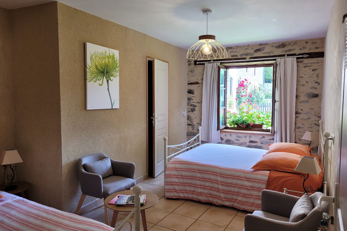 Une chambre à coucher comprenant un lit, deux chaises, une petite table, un luminaire suspendu, une fenêtre avec des rideaux et une lampe fixée au mur.