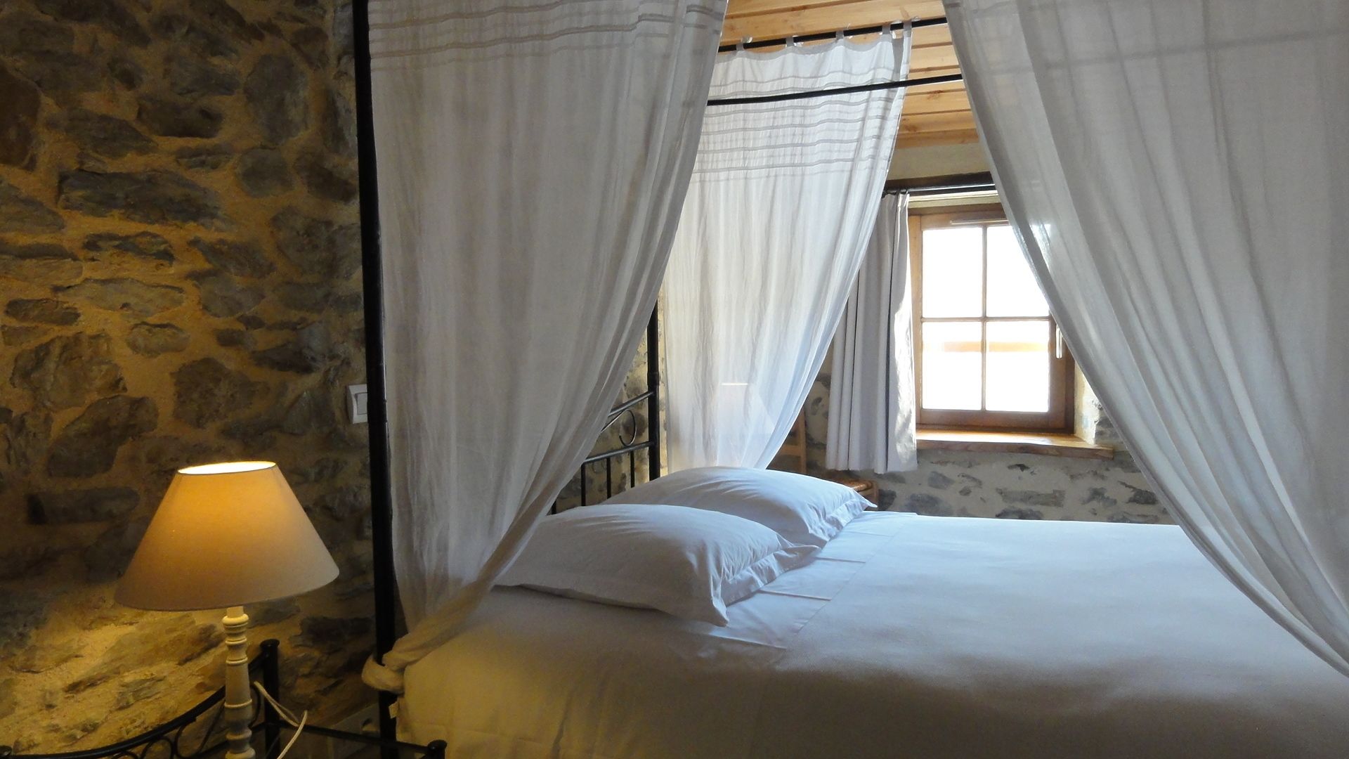 A bedroom with a stone wall, a bed with white linens, and a lamp on a bedside table.