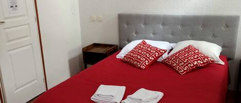 A neatly made bed with a red bedspread and two patterned pillows, a grey upholstered headboard, a small wooden nightstand, and a framed picture on the wall.