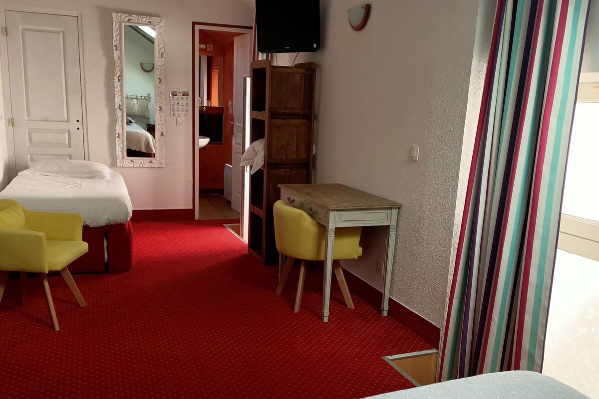 Ein Hotelzimmer mit rotem Teppich, einem Bett mit weißen Laken, einem Holztisch, einem gelben Stuhl, einem Schrank aus Holz und einem an der Wand befestigten Fernseher.