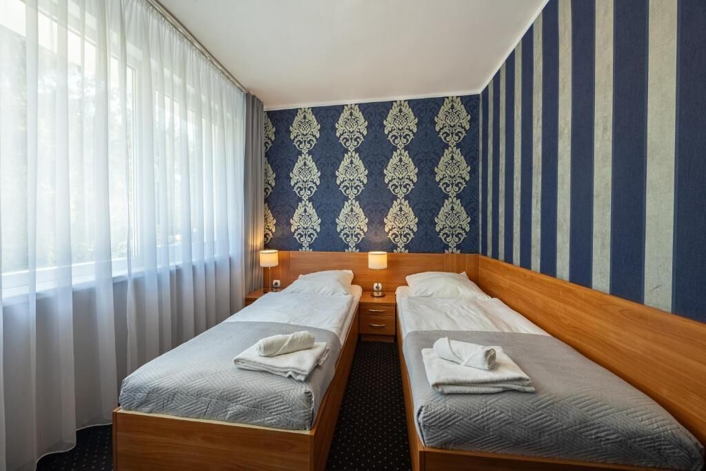 A hotel room with two single beds, a wooden headboard, a patterned wall, and a window with sheer curtains.