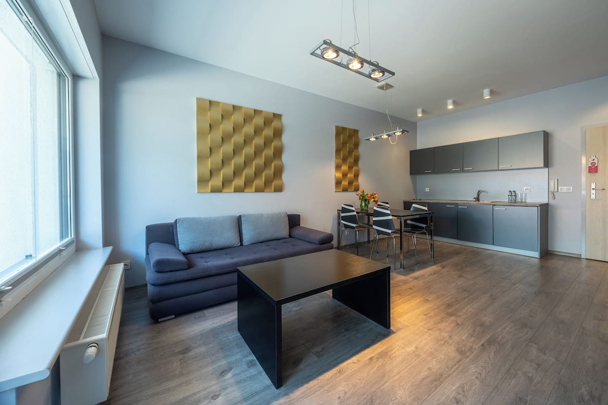 A modern living room with a sofa, a coffee table, and a dining area with chairs and a table. There is a kitchen area with cabinets and a countertop.
