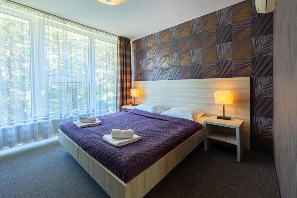 A hotel room with a large bed, two bedside tables with lamps, a window with curtains, and a patterned wall.