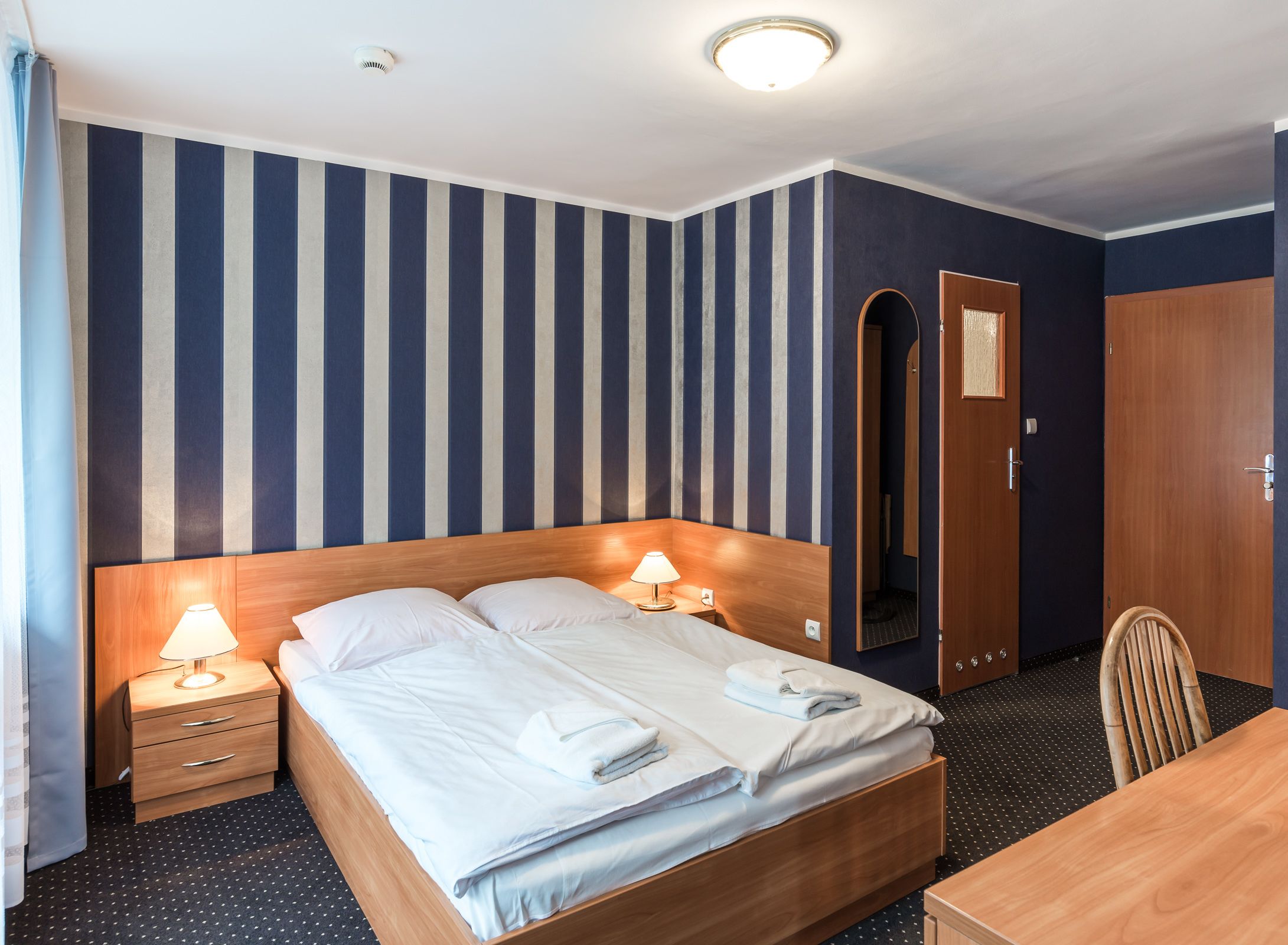 A hotel room with a large bed, two bedside tables with lamps, a wooden headboard, a wooden desk, and a wardrobe.
