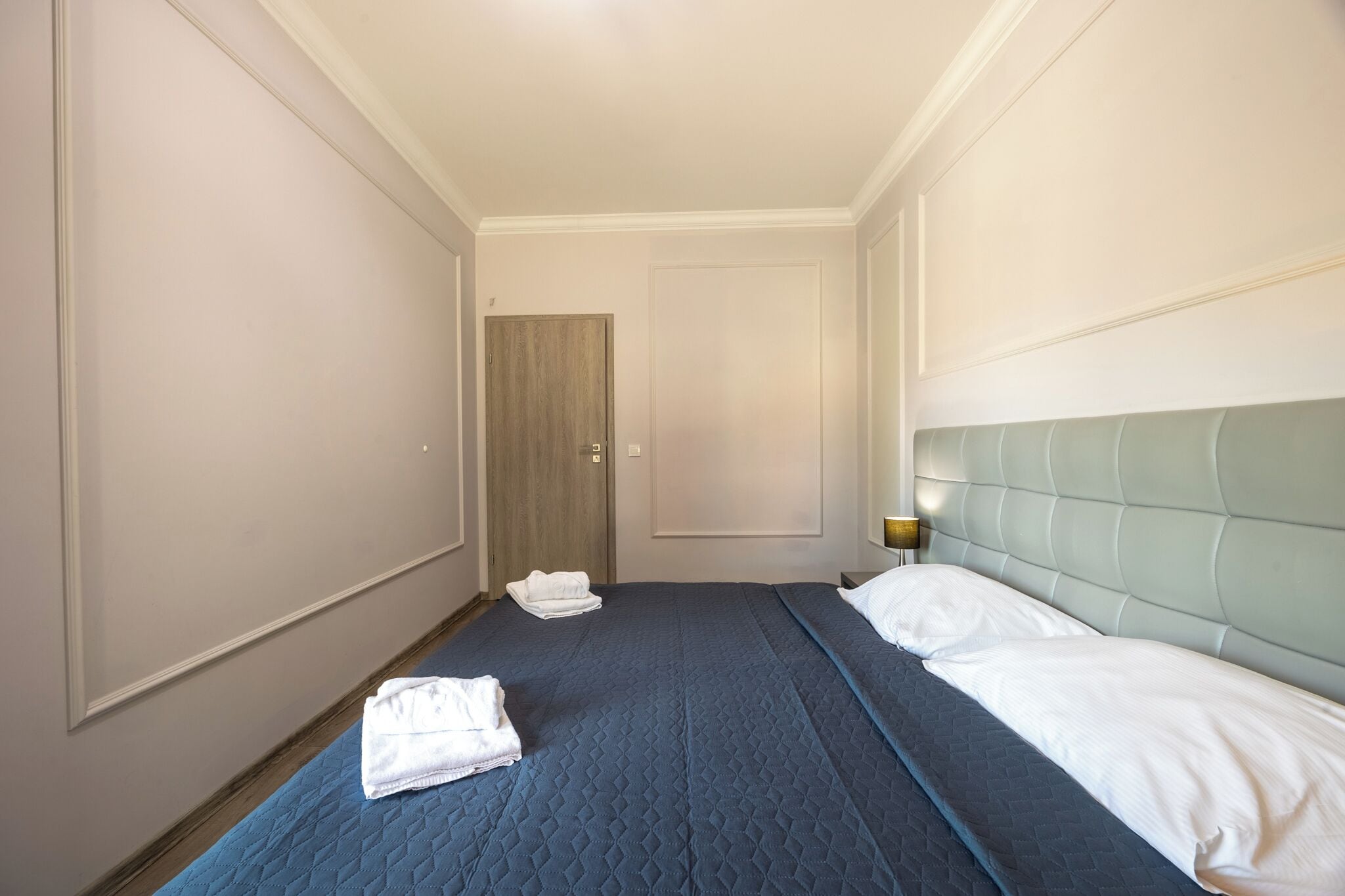 A hotel room with a single bed, a wooden door, and a nightstand with a lamp.