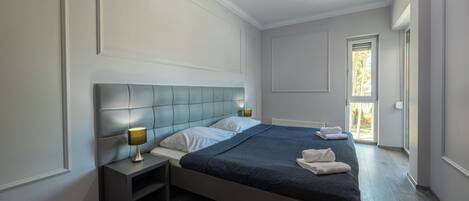 A modern bedroom with a large bed, a nightstand with a lamp, and a view of the outdoors through a window.