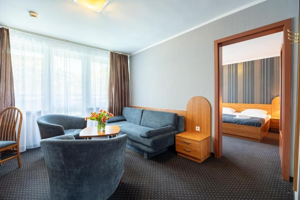 A hotel room with a sofa, a chair, a small table with flowers, a wooden nightstand, and a bed with a headboard.