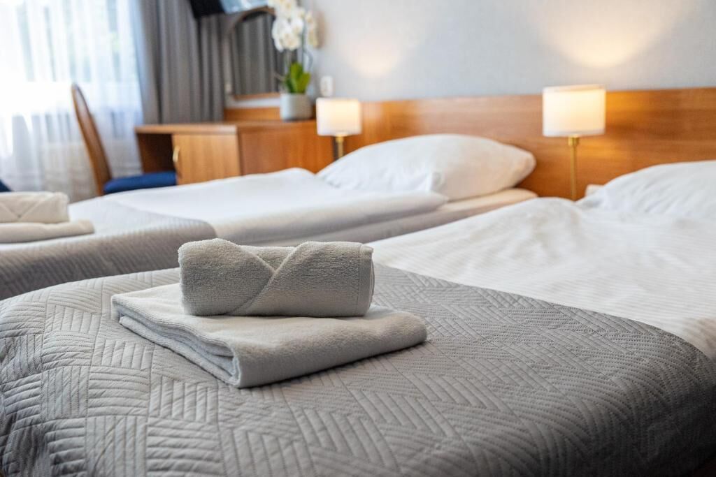 Habitación de hotel con dos camas, cada una con una toalla doblada con cuidado, un cabecero de madera, lámparas de noche y una mesita con un florero.