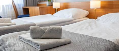 A hotel room with two beds, each with a neatly folded towel, a wooden headboard, bedside lamps, and a small table with a flower vase.