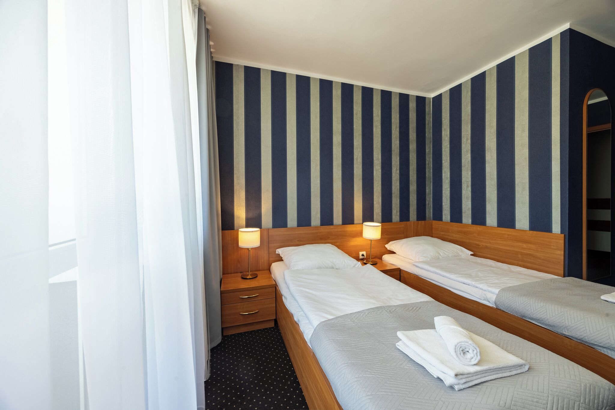 A hotel room with two single beds, wooden headboards, bedside tables with lamps, and striped wallpaper.