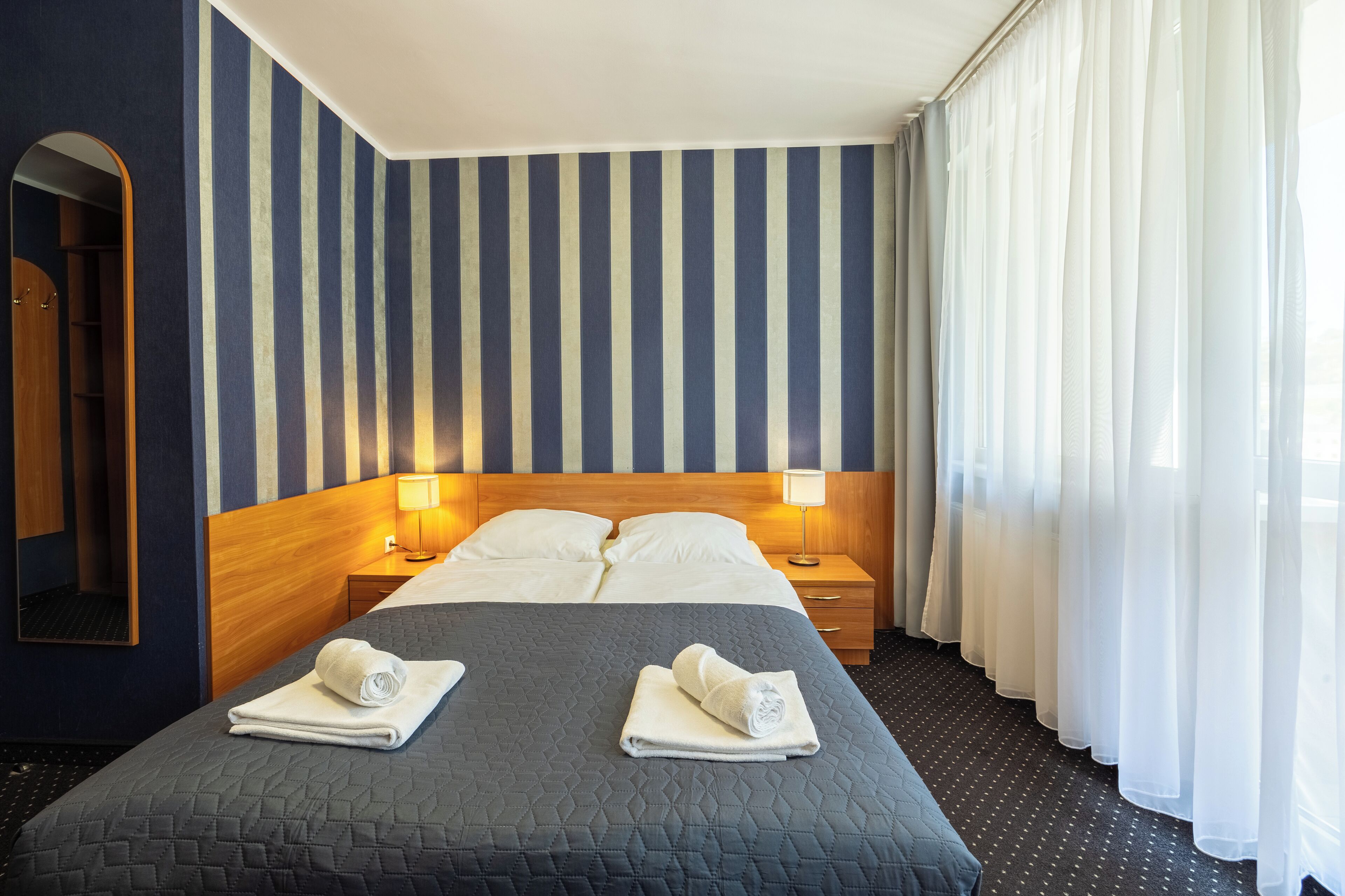 A hotel room with a large bed, two bedside tables with lamps, a wooden headboard, a mirror, and a window with sheer curtains.