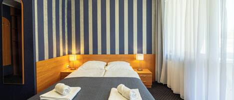 A hotel room with a large bed, two bedside tables with lamps, a wooden headboard, a mirror, and a window with sheer curtains.