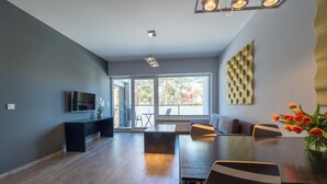 A modern living room with a dining area, large windows, and a flat-screen TV on a black stand.